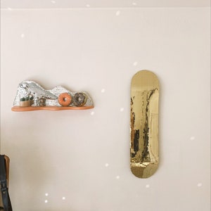 Puede incluir: Un espejo dorado con forma de tabla de skate cuelga de una pared blanca con un peque&ntilde;o estante debajo. El estante est&aacute; decorado con una escultura de mosaico plateado y blanco, dos peque&ntilde;as plantas en maceta y dos rosquillas naranjas.
