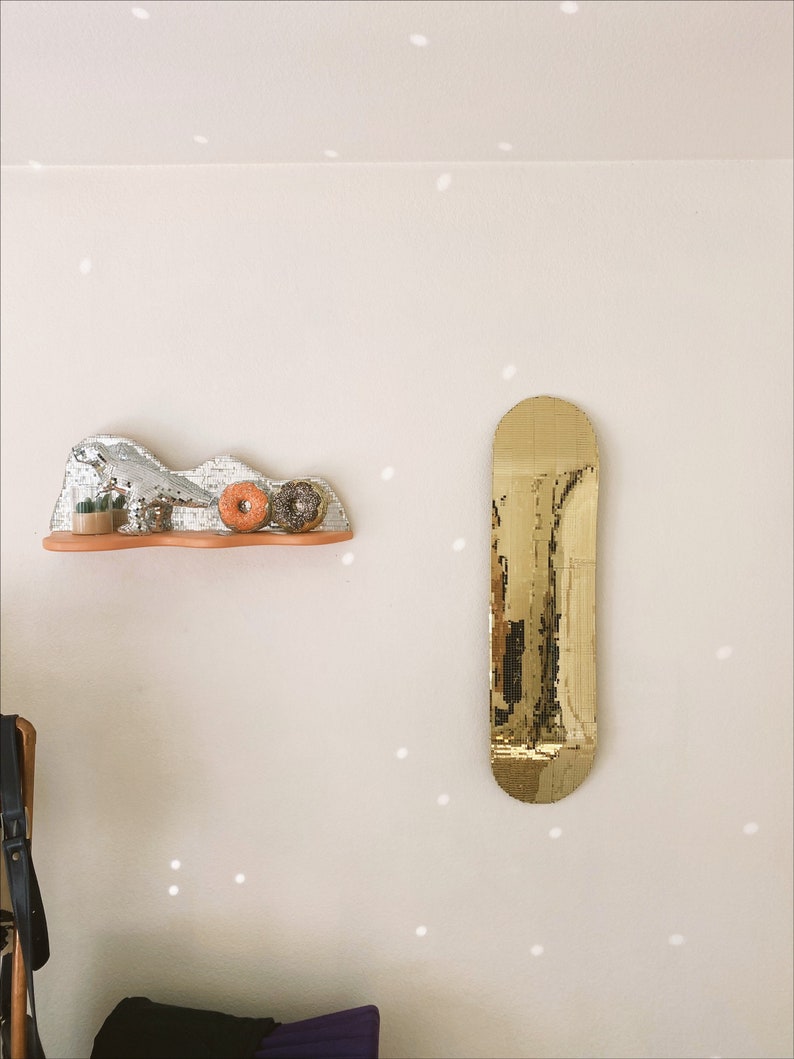 Puede incluir: Un espejo dorado con forma de tabla de skate cuelga de una pared blanca con un peque&ntilde;o estante debajo. El estante est&aacute; decorado con una escultura de mosaico plateado y blanco, dos peque&ntilde;as plantas en maceta y dos rosquillas naranjas.