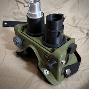 以下が含まれることがあります： オリーブグリーンの双眼鏡型デバイス。2つの黒い円筒形のレンズハウジングを備え、片方には銀色のトップアタッチメントが付いています。側面ダイヤル、黒いストラップ、PULL IR、ON、OFFと記されたコントロールパネルが装備されています。