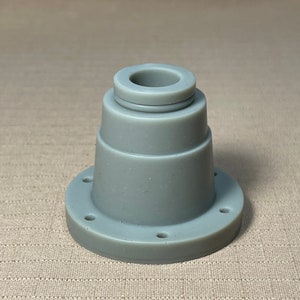 Op de afbeelding: Een grijs, cilindrisch object met een brede basis en een kleinere, cirkelvormige bovenkant. De basis heeft verschillende gelijkmatig verdeelde gaten. Het object lijkt gemaakt van een solide, glad materiaal. De vorm suggereert een mechanisch of industrieel onderdeel.