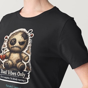 May include: Black t-shirt featuring a distressed voodoo doll graphic with the text "Bad Vibes Only." The doll has a stitched appearance, a button eye, and pins. The text "If it's negative, I'm already expecting it" is also included.