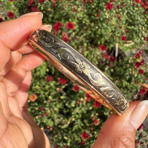 Peut inclure: Un bracelet jonc en métal doré orné d'un motif gravé de fleurs et de volutes. La surface du bracelet présente un fond texturé et assombri dans les zones en creux du motif, ce qui met en valeur les détails floraux métalliques en relief.