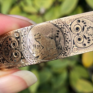 Pode incluir: Um bracelete dourado com um design gravado que apresenta um sapo em um círculo rodeado de motivos florais.