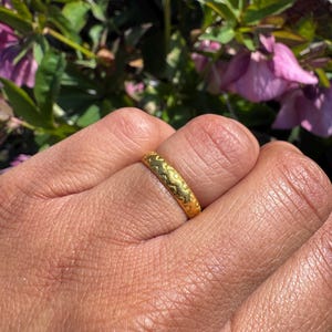 May include: A gold ring featuring a repeating pattern of curved lines. The ring is on a finger, with a blurred background of green leaves and pink flowers.