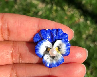 May include: A detailed blue and white pansy flower with a pearl centre. The flower has a glossy finish. The background is blurred, suggesting an outdoor setting. The flower is a decorative item.