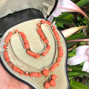 Puede incluir: Un conjunto de collar y pendientes de coral vintage en una caja de joyería de terciopelo negro. El collar está hecho de cuentas de coral talladas y cadena de oro. Los pendientes son pendientes colgantes con formas de lágrima de coral.