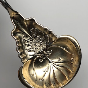 May include: A silver serving spoon with an ornate, gold-plated bowl. The bowl is decorated with a floral design and has a scalloped edge.