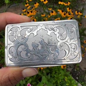 May include: A rectangular silver-toned metal case featuring an intricate engraved design on the lid. The center depicts a figure standing near a large bowl and a pagoda structure, surrounded by elaborate scrolling patterns and fine line detailing.