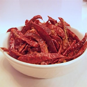 May include: A white bowl filled with dried red chili peppers. The peppers are a deep red color and have a wrinkled texture. The bowl is round and sits on a white surface.
