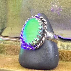 May include: A silver ring featuring a bright, glowing green oval stone set within a decorative, scalloped metal bezel and a twisted rope-style border. The ring is displayed on a dark, smooth, triangular stand, showcasing its vibrant colour.