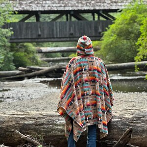 Puede incluir: Una persona con un poncho con capucha y un patr&oacute;n geom&eacute;trico colorido se encuentra cerca de un puente de madera sobre un arroyo. El poncho es marr&oacute;n, naranja, rojo, azul y blanco. La persona lleva pantalones vaqueros azules.