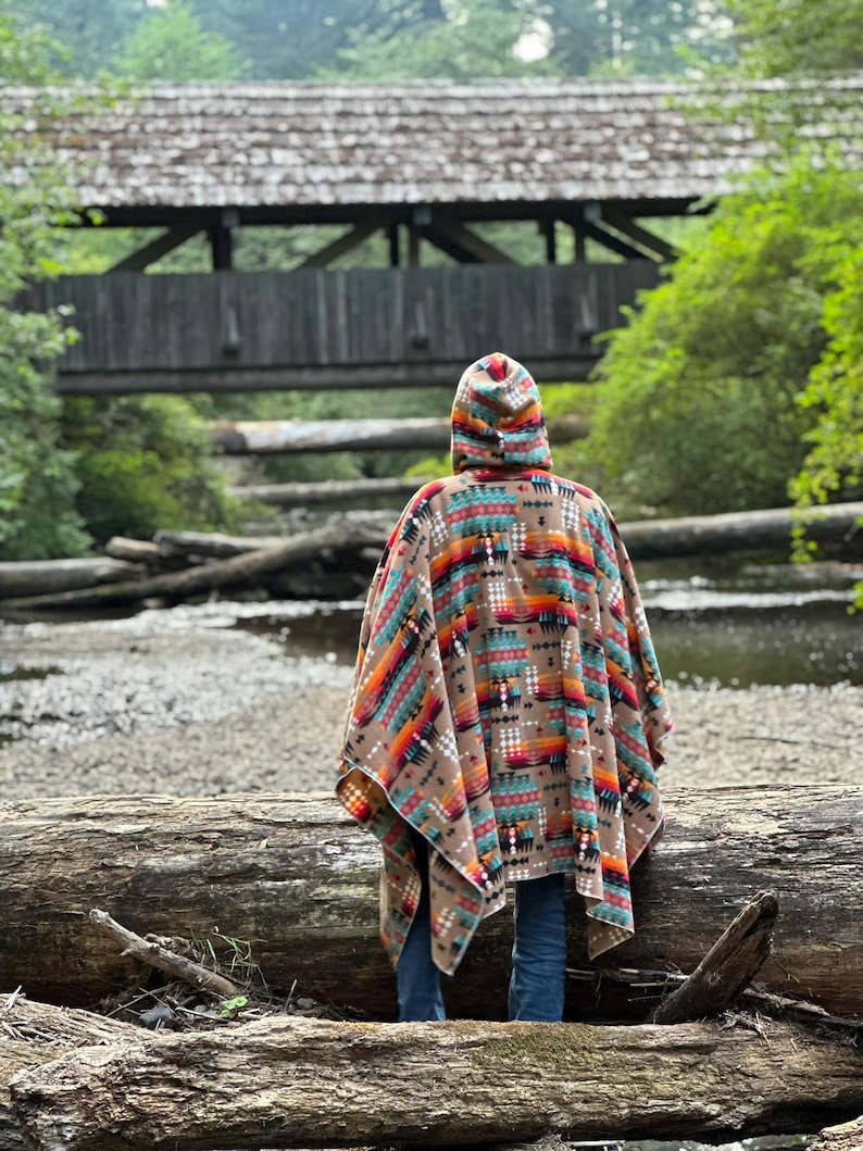 Puede incluir: Una persona con un poncho con capucha y un patr&oacute;n geom&eacute;trico colorido se encuentra cerca de un puente de madera sobre un arroyo. El poncho es marr&oacute;n, naranja, rojo, azul y blanco. La persona lleva pantalones vaqueros azules.