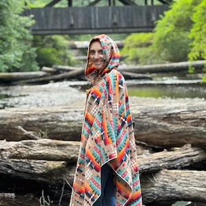 Puede incluir: Una mujer lleva una capucha con un poncho con un patr&oacute;n geom&eacute;trico colorido. El poncho es marr&oacute;n, naranja, rojo, azul y blanco. Ella est&aacute; de pie frente a un puente de madera sobre un arroyo.