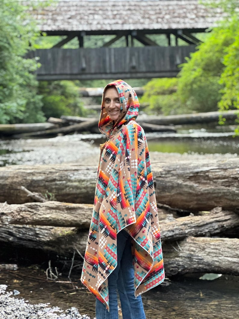 Puede incluir: Una mujer lleva una capucha con un poncho con un patr&oacute;n geom&eacute;trico colorido. El poncho es marr&oacute;n, naranja, rojo, azul y blanco. Ella est&aacute; de pie frente a un puente de madera sobre un arroyo.