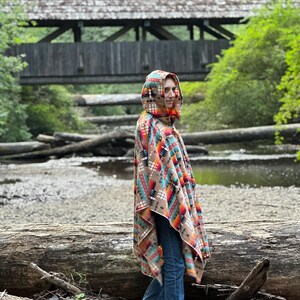 Puede incluir: Una persona lleva una capucha con un patr&oacute;n geom&eacute;trico colorido. La capucha es marr&oacute;n, naranja, roja, azul y blanca. La persona est&aacute; de pie en un entorno boscoso con un puente de madera en el fondo.