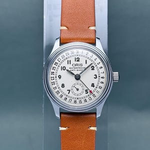 May include: A vintage ORIS wristwatch with a cream-colored face, silver case, and brown leather band. The watch face features black numerals, a subdial, and the text "ORIS WATERPROOF ANTI-SHOCK". The band is secured with white stitching.