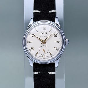 May include: A vintage Oris wristwatch with a silver-tone stainless steel case and a cream-colored dial. The dial features gold-tone Arabic numerals at 2, 4, 8, 10, and 12 o'clock, gold-tone hands, a sub-dial for seconds, and a black suede leather strap.