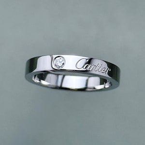 May include: A polished silver-toned metal band ring featuring a single round brilliant-cut clear gemstone set flush into the surface. The word Cartier is engraved in a cursive script on the outer face of the band next to the gemstone setting.
