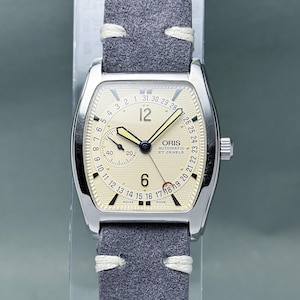 May include: A silver wristwatch with a cream face and a grey suede strap. The watch face displays the brand "ORIS", along with the date and time. The watch has a rectangular shape with a curved top and bottom. The strap is approximately 20 cm long.