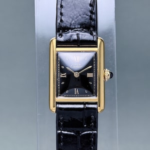 May include: A rectangular wristwatch with a gold-tone case and a black dial featuring the text Must de Cartier. The watch has gold-tone hands, Roman numeral markers at the twelve, three, six, and nine o'clock positions, and a black leather strap.