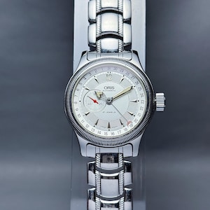 Może przedstawiać: Srebrny zegarek automatyczny Oris z białą tarczą i srebrnym metalowym paskiem. Zegarek ma datownik w okienku na godzinie 3 i 24-godzinny wyświetlacz wokół zewnętrznej krawędzi tarczy. Tarcza zegarka zawiera również tekst "ORIS AUTOMATIC" i "27 JEWELS".