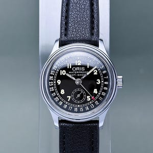 Peut inclure: Une montre-bracelet argentée avec un cadran noir, des chiffres arabes blancs et un bracelet en cuir noir. Le cadran porte les inscriptions ORIS, WATERPROOF et ANTI-SHOCK, avec un anneau de date numéroté de 1 à 31 et une aiguille à pointe rouge.
