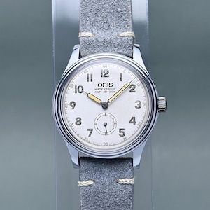 May include: A silver-toned wristwatch with a round white dial, featuring black Arabic numerals, gold-toned hour and minute hands, and a small seconds sub-dial at the six o'clock position. The dial displays the text ORIS WATERPROOF ANTI-SHOCK.