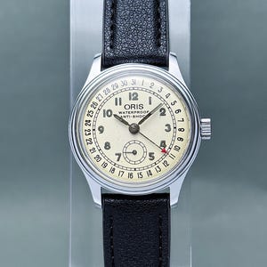 Peut inclure: Une montre-bracelet Oris vintage avec un cadran crème, des chiffres arabes noirs et une aiguille de date avec une pointe de flèche rouge. La montre présente un boîtier rond argenté, un bracelet en cuir noir et l'inscription ORIS WATERPROOF ANTI-SHOC.