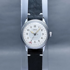 May include: A silver Oris wristwatch with a white face and black leather strap. The watch has a date window and is water resistant. The watch face has the text "ORIS 17 JEWELS WATERPROOF ANTISHOCK" printed on it.