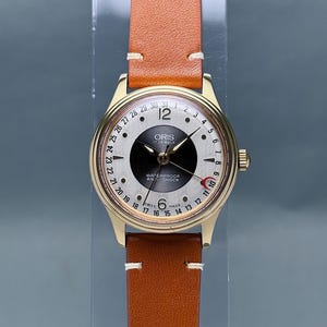 Może przedstawiać: Złoty zegarek na rękę z brązowym skórzanym paskiem. Tarcza zegarka jest biało-czarna z napisem "ORIS" i "WATERPROOF ANTI-SHOCK". Zegarek posiada wyświetlacz daty i wskazówkę sekundową z czerwoną końcówką.