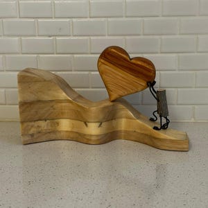 Puede incluir: Una escultura de madera con un elemento en forma de corazón sostenido por una pequeña figura. El corazón es de color marrón cálido, y la base es de madera clara con un diseño ondulado. La figura parece estar hecha de madera y alambre.