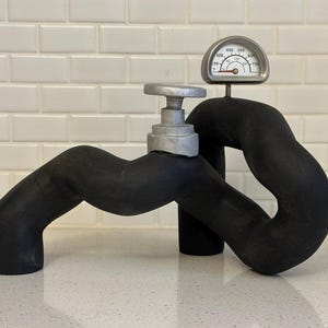 May include: A black rubber pipe sculpture with a silver valve and a round gauge that reads from 0 to 700. The gauge has a red needle pointing to 500.