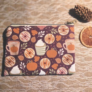 Peut inclure: Une pochette rectangulaire à fermeture éclair avec un fond violet foncé orné d'un motif répétitif de citrouilles orange, de beignets au glaçage blanc, de tasses de café décorées de citrouilles et de feuilles d'automne brunes et crème.