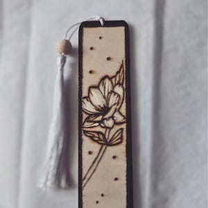 May include: A wooden bookmark with a floral design. The bookmark has a light brown background and a dark brown border. A flower and leaves are etched into the wood. A white tassel and wooden bead are attached to the top.