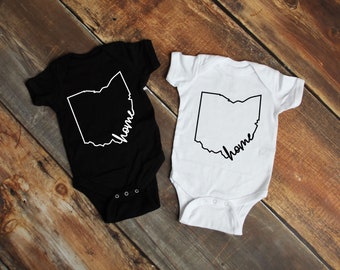 May include: Two white baby bodysuits with short sleeves. One bodysuit is black and the other is white. Both bodysuits have a black outline of the state of Ohio with the word "home" written inside the outline.
