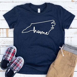 May include: A navy blue t-shirt with a white outline of the state of North Carolina and the word "home" written inside the outline. The shirt is laying on a white wooden surface with a pair of red and black plaid shoes and tan shorts.