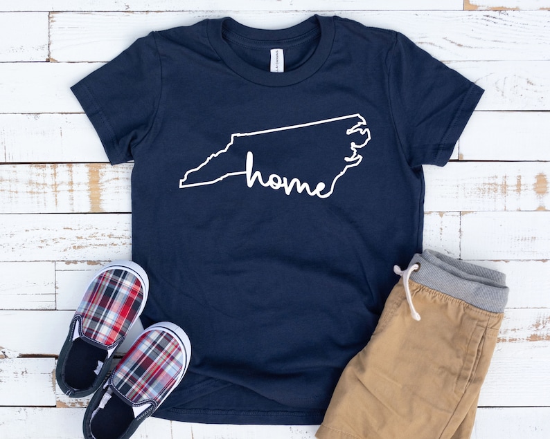 May include: A navy blue t-shirt with a white outline of the state of North Carolina and the word "home" written inside the outline. The shirt is laying on a white wooden surface with a pair of red and black plaid shoes and tan shorts.