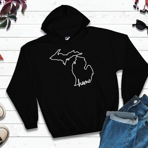 May include: Black hooded sweatshirt featuring a white outline of the state of Michigan with the word "home" in a cursive font. The sweatshirt is displayed with a pair of blue jeans and sneakers.