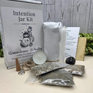 May include: An Intention Jar Kit with a white box featuring text and illustrations. The kit includes a candle, incense cone, small bags of herbs, a black stone, and a scroll. The kit is designed for remembrance and healing.