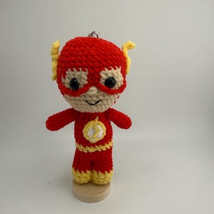 May include: A handmade crochet Flash superhero doll. The doll is primarily red with yellow accents, including the lightning bolt emblem on the chest and the mask. The doll is standing on a small wooden base.