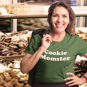 May include: A woman wearing a green t-shirt with the text "Cookie Momster" in white letters. She is standing in front of a bakery display case filled with various pastries and cookies.