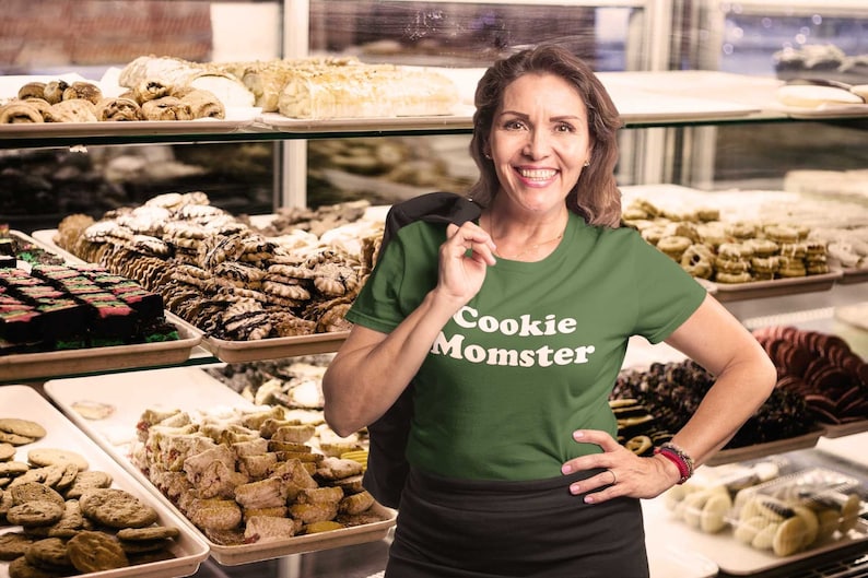 May include: A woman wearing a green t-shirt with the text "Cookie Momster" in white letters. She is standing in front of a bakery display case filled with various pastries and cookies.