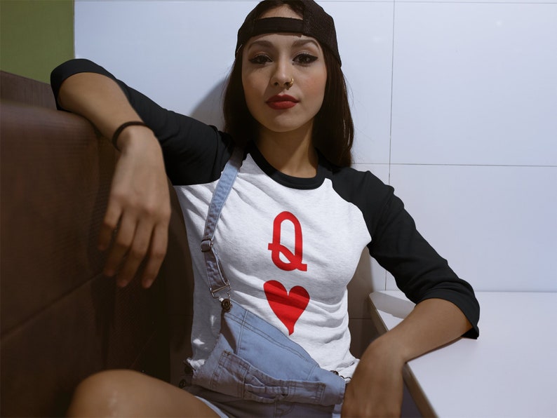 Op de afbeelding: Een vrouw draagt een wit en zwart baseballshirt met een rode grafische afbeelding van de hartenkoningin. Ze draagt een zwarte baseballpet en een blauwe denim overall.
