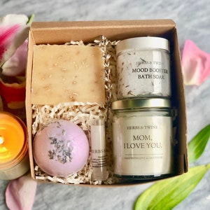 May include: A gift box filled with bath and body products. The box contains a lavender bath bomb, a candle with the text "Mom, I love you." a bar of soap, and a jar of bath salts. The text on the jar of bath salts reads "Mood Booster Bath Soak".