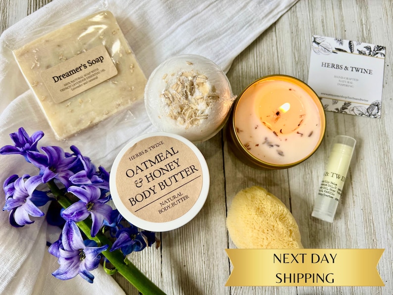 May include: A collection of self-care products including a bar of Dreamer's Soap, a bath bomb, a candle, body butter, lip balm, and a natural sponge. The products are arranged with purple hyacinth flowers on a white cloth.