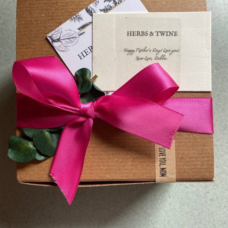 May include: A brown cardboard gift box with a pink satin ribbon tied in a bow. The box has a small white tag with the text "HERBS & TWINE" and the message "Happy Mother's Day! Love you! Your Love, Debbie". The box also has a brown paper tag with the text "LOVE YOU, MOM".