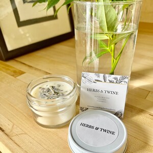 May include: A white candle in a clear glass jar with a white lid. The candle has dried herbs and flowers embedded in it. The jar has a white label that says "Herbs & Twine" and "Hand-crafted, Natural, Inspiring". The website address is also included on the label: "www.etsy.com/shop/herbsandtwine".