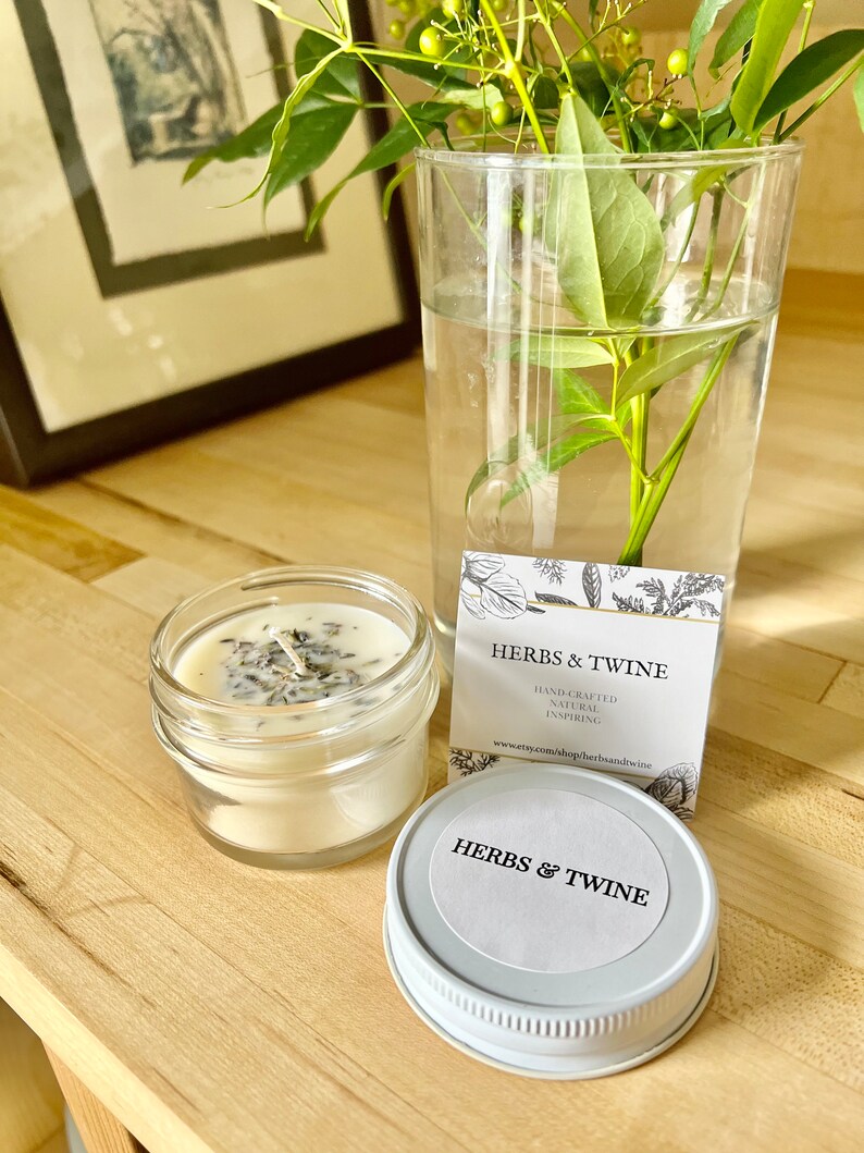 May include: A white candle in a clear glass jar with a white lid. The candle has dried herbs and flowers embedded in it. The jar has a white label that says "Herbs & Twine" and "Hand-crafted, Natural, Inspiring". The website address is also included on the label: "www.etsy.com/shop/herbsandtwine".