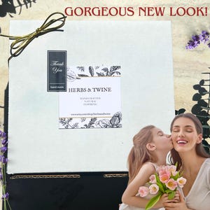 May include: A white box with a gold ribbon, a label that reads "HERBS & TWINE", and the text "GORGEOUS NEW LOOK!". A woman and a child are in the foreground, with the child kissing the woman's cheek. The woman is holding a bouquet of pink flowers.