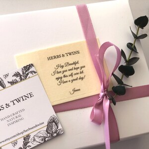 May include: A white gift box with a pink ribbon and a small sprig of eucalyptus. The box is tied with a pink ribbon and has a small card attached to it that says "HERBS & TWINE" and "Hey Beautiful, I love you and hope you enjoy this self care kit. Have a great day! - Jenna".